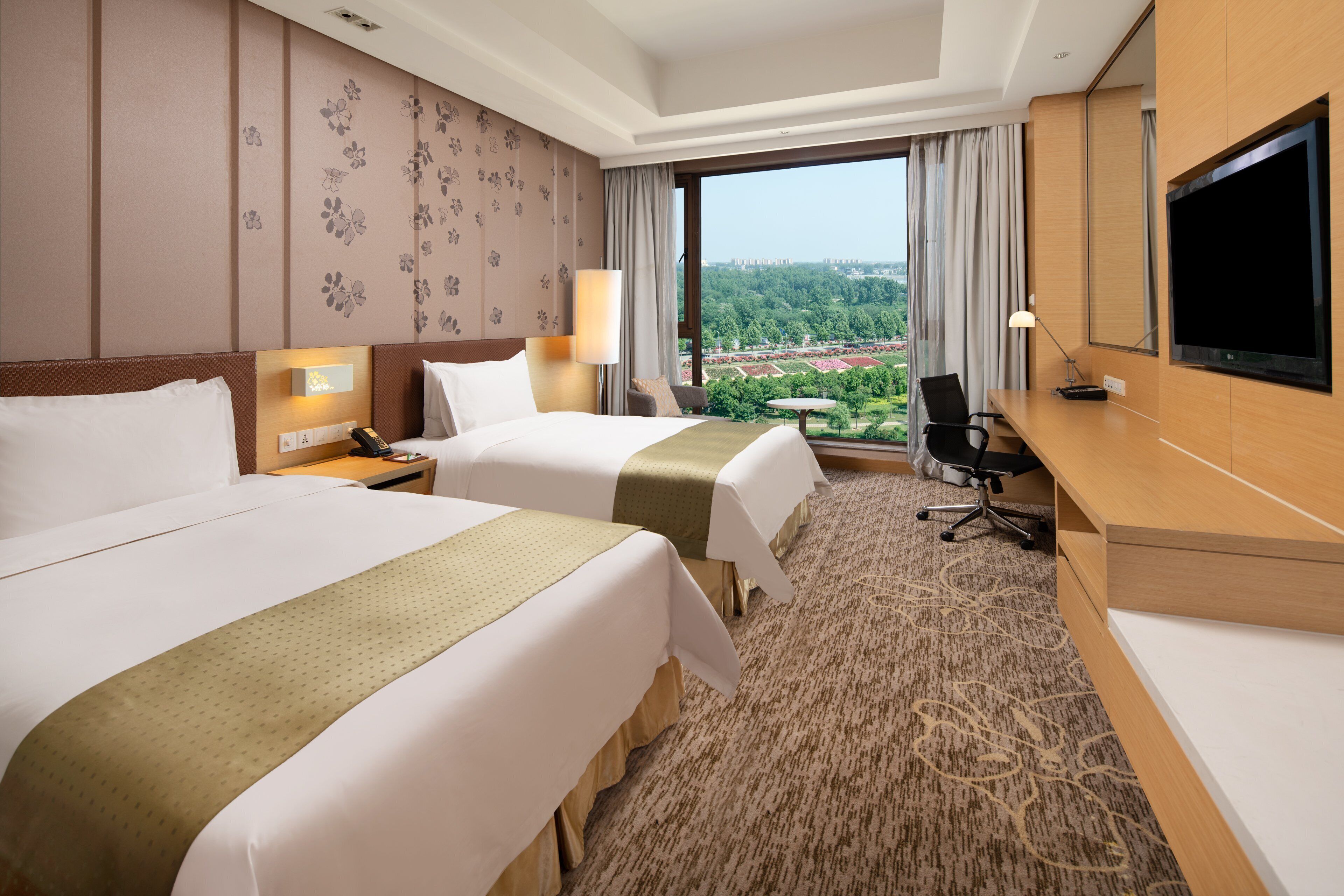 A hotel room with two beds, a desk, a TV, and a view of a landscaped area.