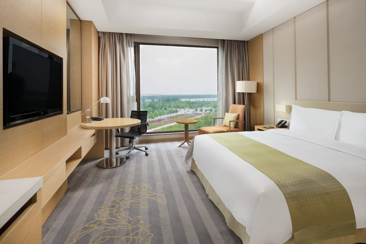 Quarto de hotel com uma cama grande, uma escrivaninha com cadeira, uma televisão e vista para a paisagem urbana através de uma janela ampla.