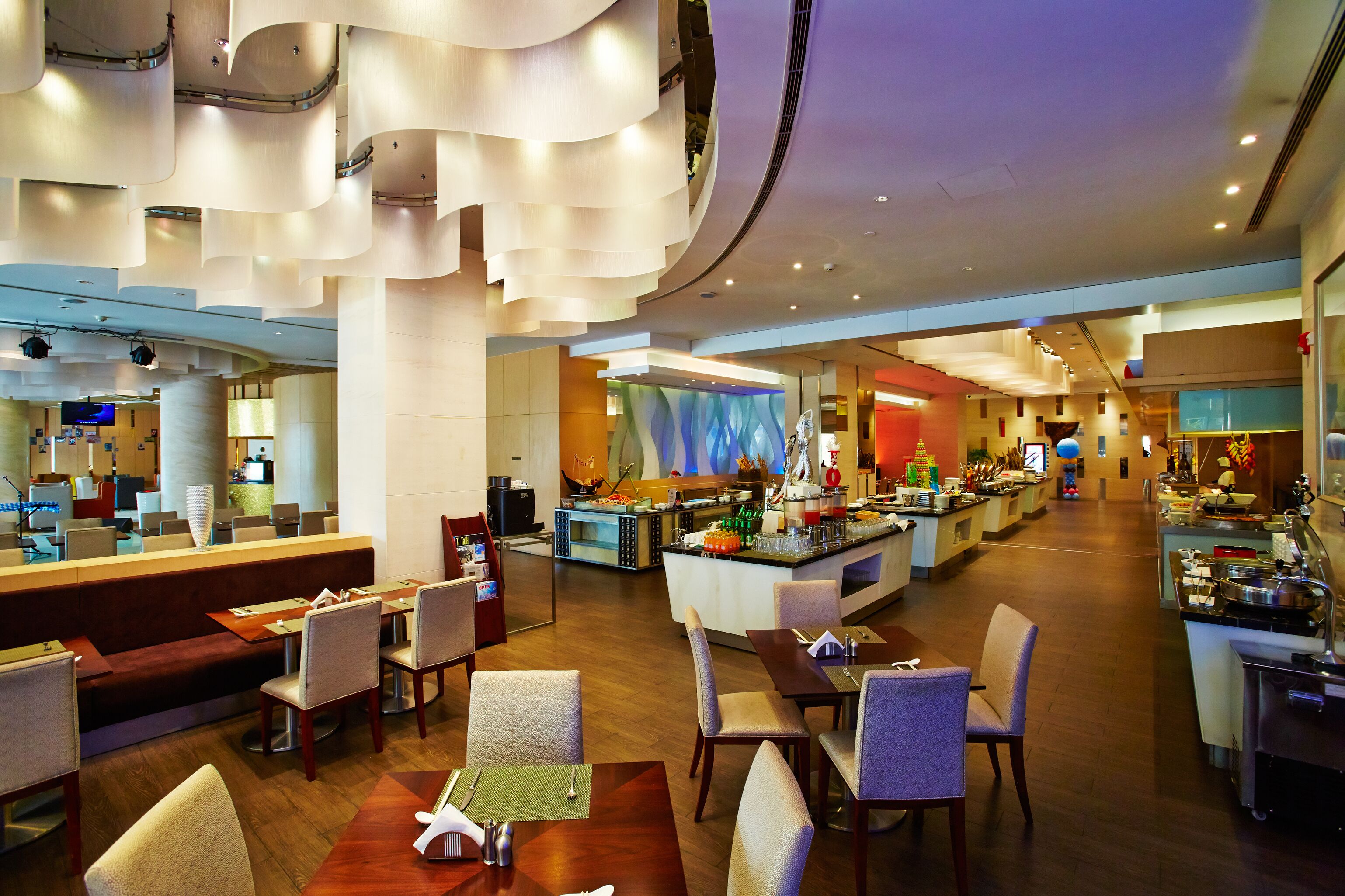 Een moderne hotellobby met een centraal gelegen bar, zithoekjes en zicht op de keuken op de achtergrond.
