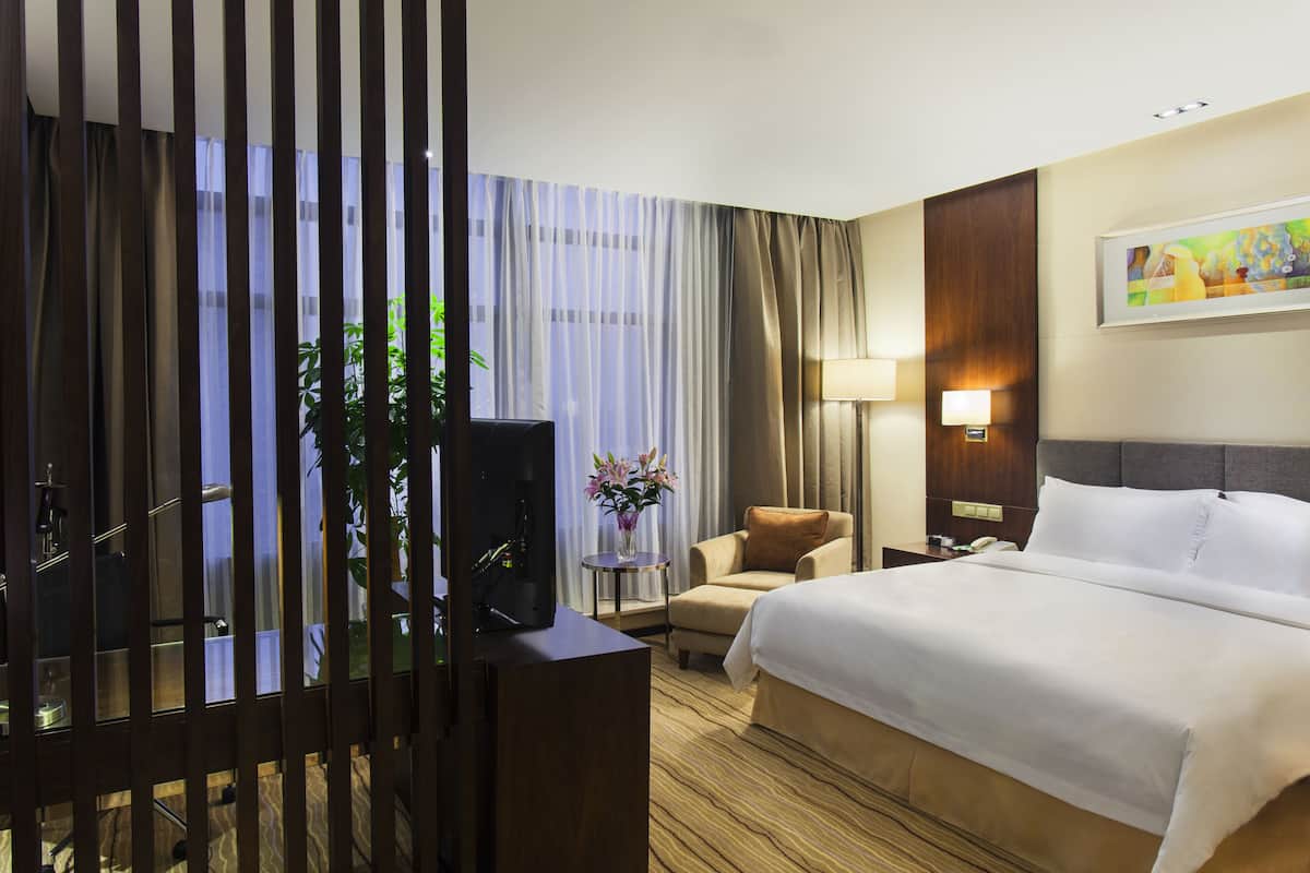 A hotel room with a bed, a chair, a desk with a computer, a lamp, and a painting on the wall.
