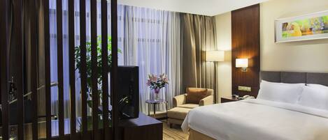 A hotel room with a bed, a chair, a desk with a computer, a lamp, and a painting on the wall.