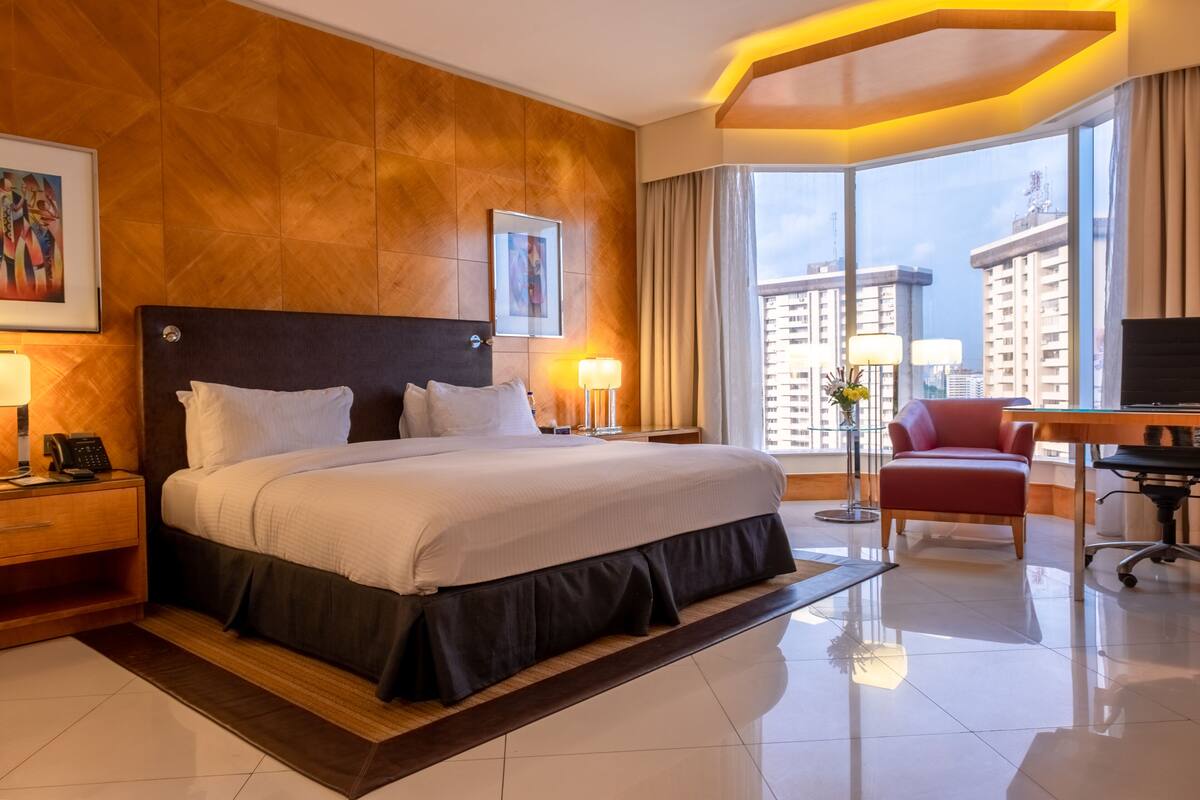 A modern hotel room with a large bed, a desk, and a view of high-rise buildings.