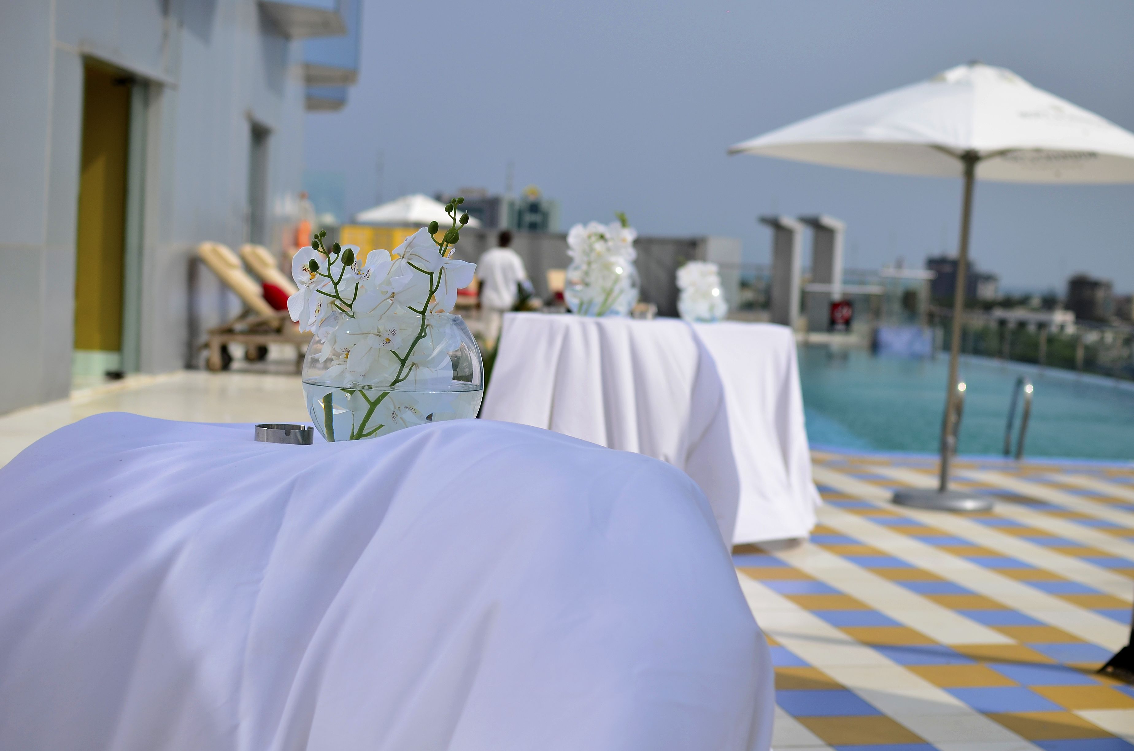 A poolside area with white tablecloths and flower arrangements, a pool, and a cityscape in the background.