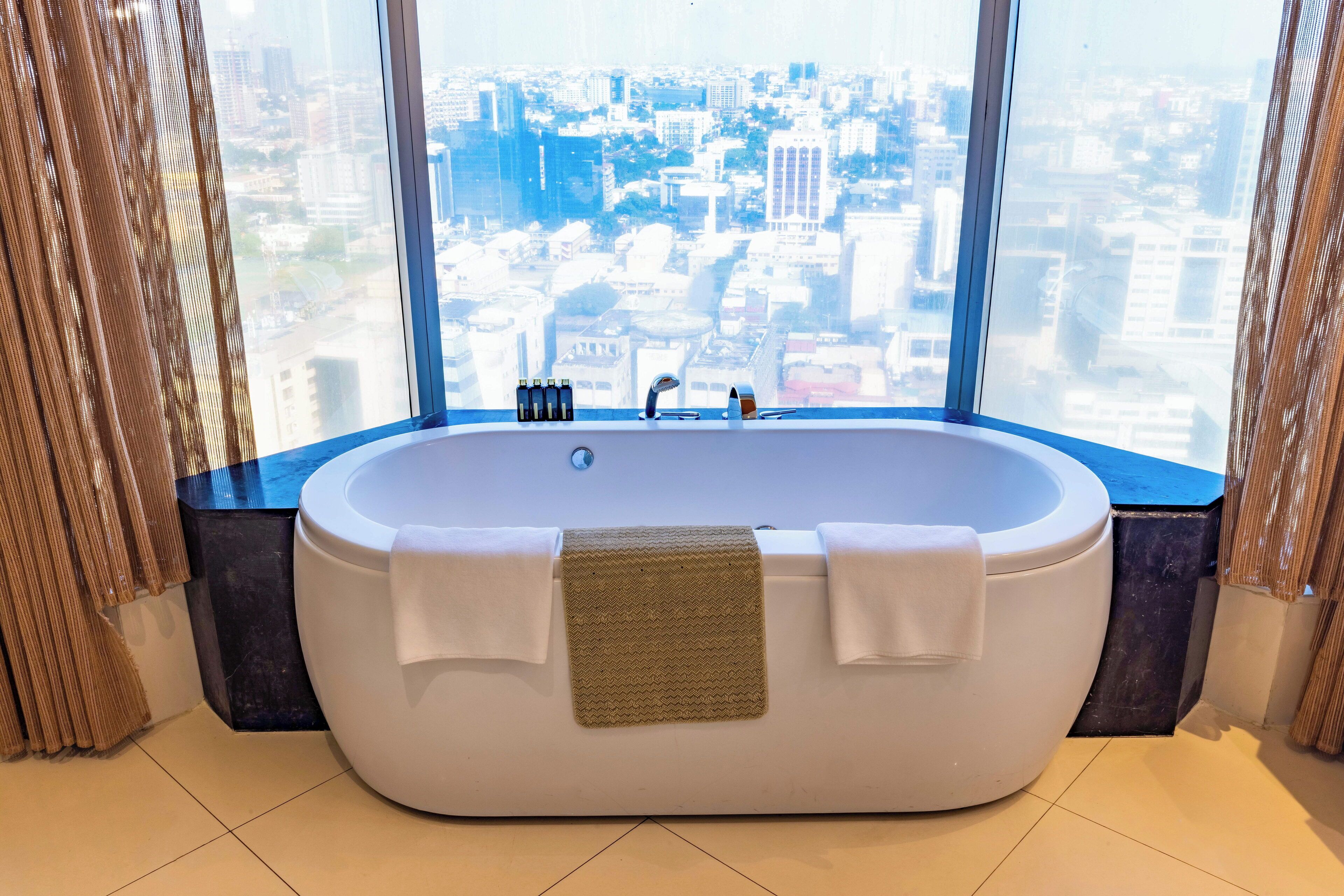 A modern bathroom with a freestanding bathtub, a view of a cityscape through large windows, and a towel rack with towels.
