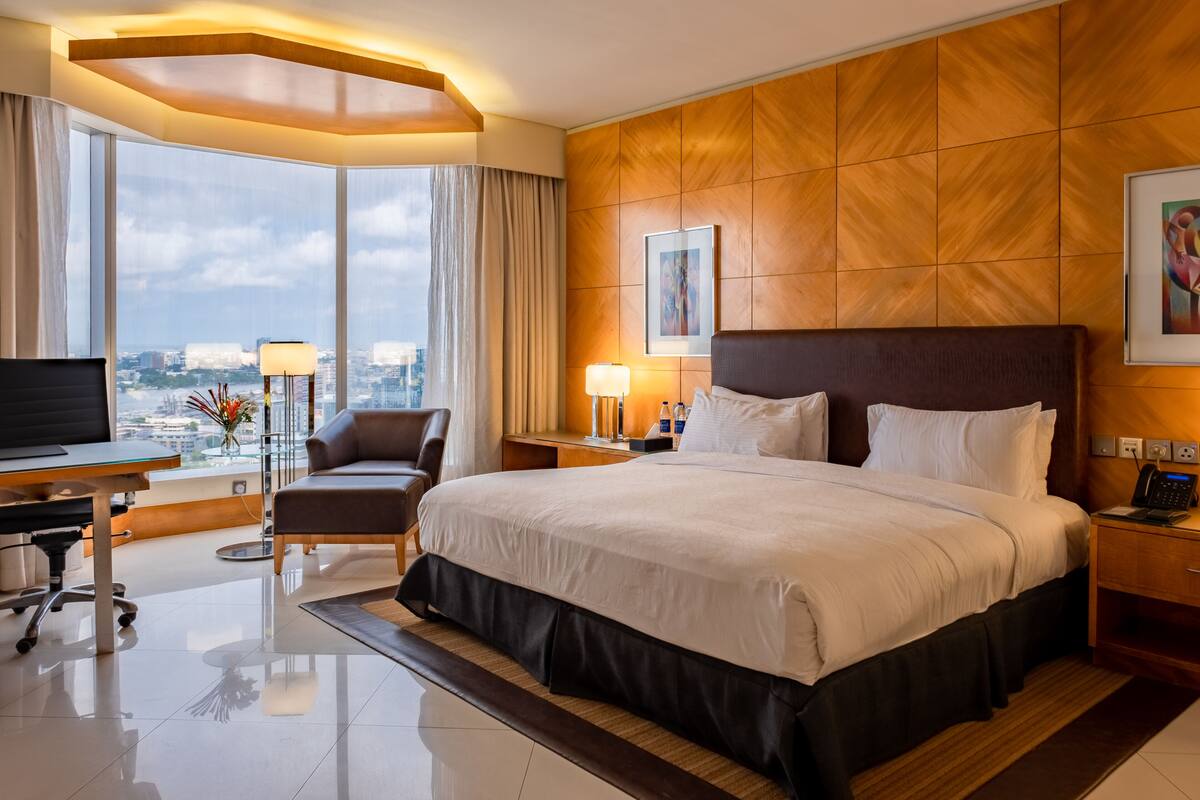 A hotel room with a large bed, a desk with a computer, a chair, a TV, and a view of the city through large windows.