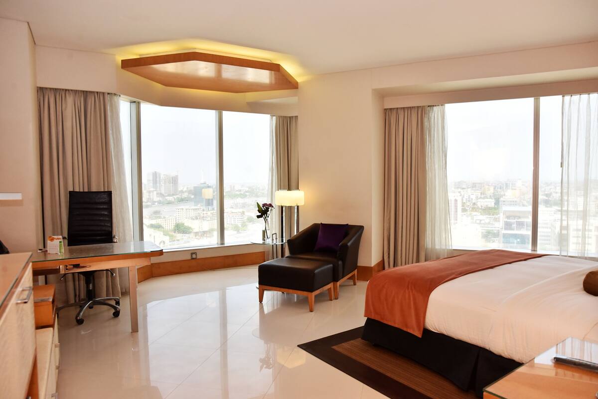 A hotel room with a bed, desk, chair, and large windows offering a city view.