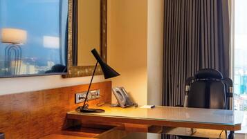 A hotel room with a wooden desk, a mirror, a lamp, a chair, and a view of the city through the window.