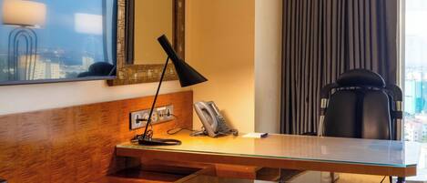 Habitación de hotel con escritorio de madera, espejo, lámpara, silla y vista a la ciudad por la ventana.