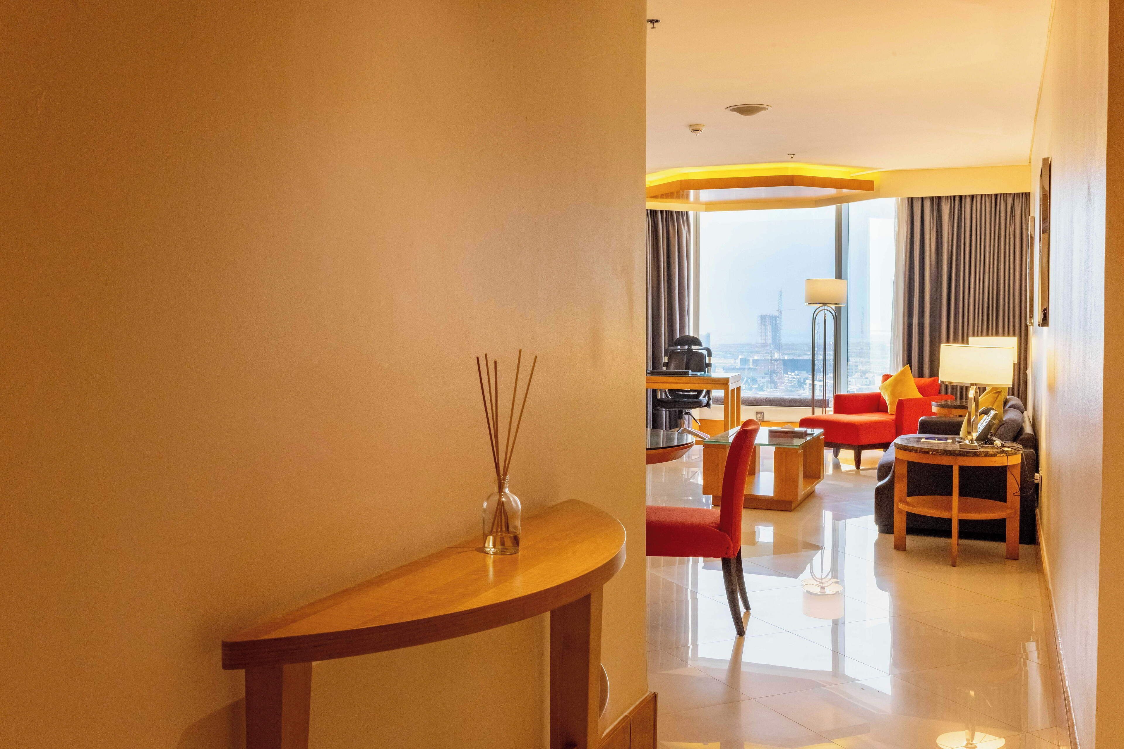 A hotel room with a wooden console table, a red chair, a sofa, a coffee table, and a lamp.