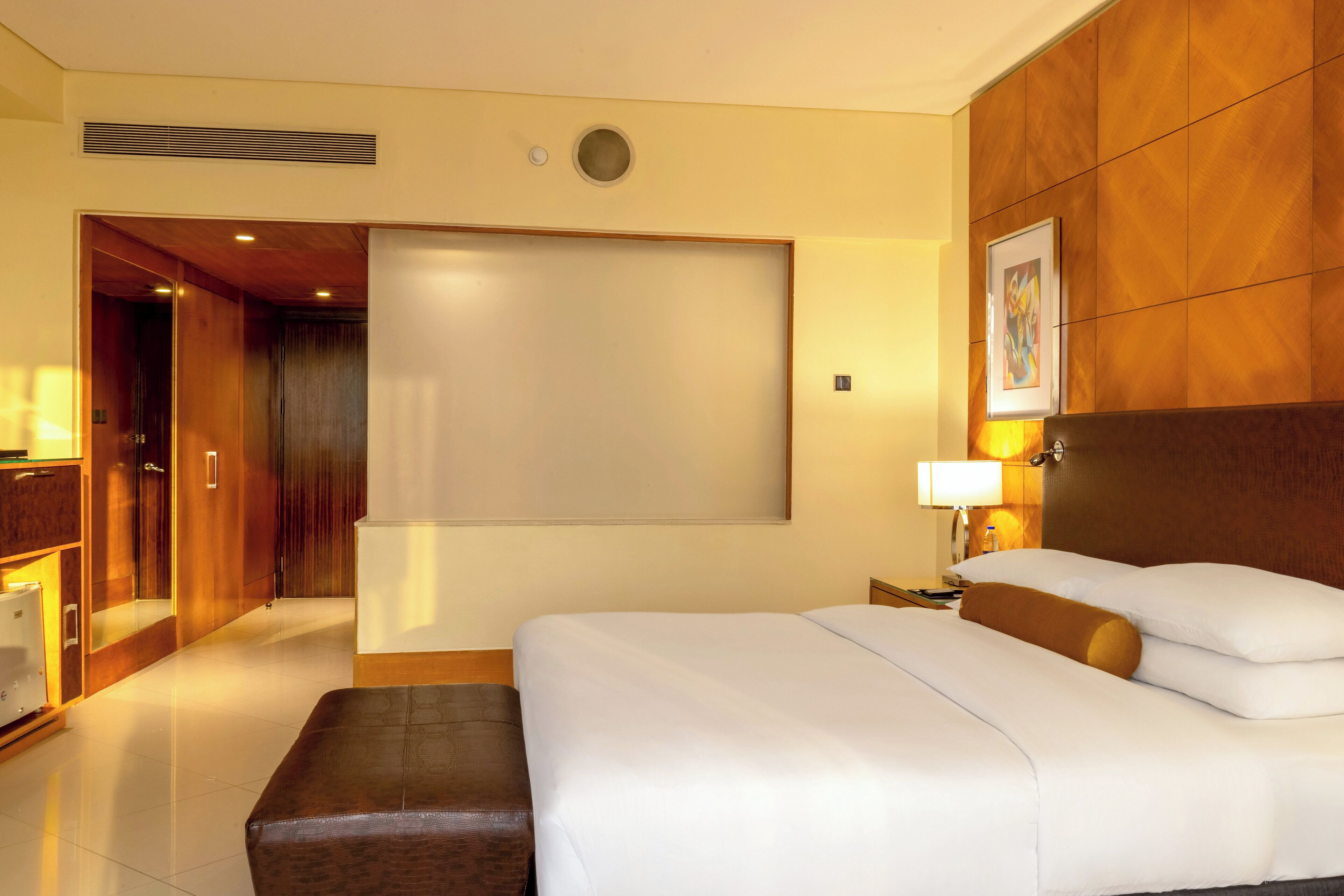 A hotel room with a bed, a bedside table with a lamp, a wooden headboard, a large window, and a door leading to a bathroom.