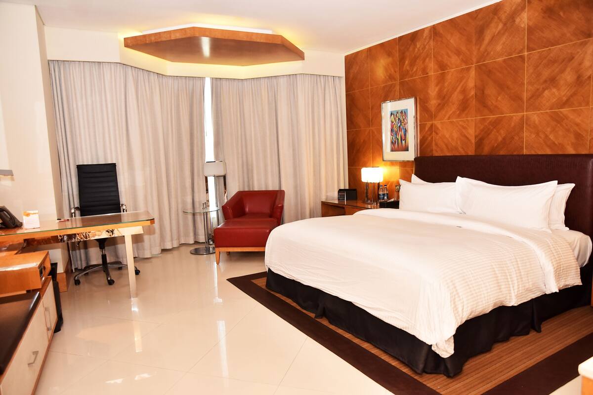 A hotel room with a large bed, a desk, a red chair, a TV, and a painting on the wall.