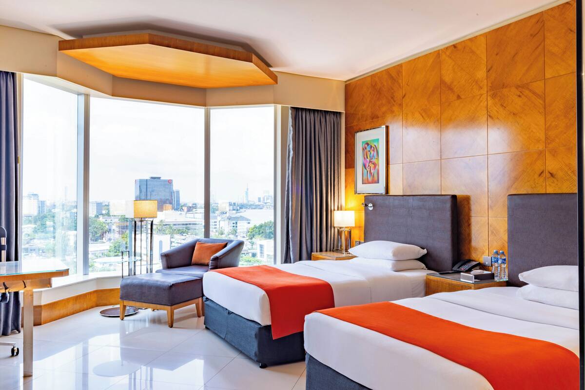 A hotel room with two beds, a sofa, a desk, and a large window offering a city view.