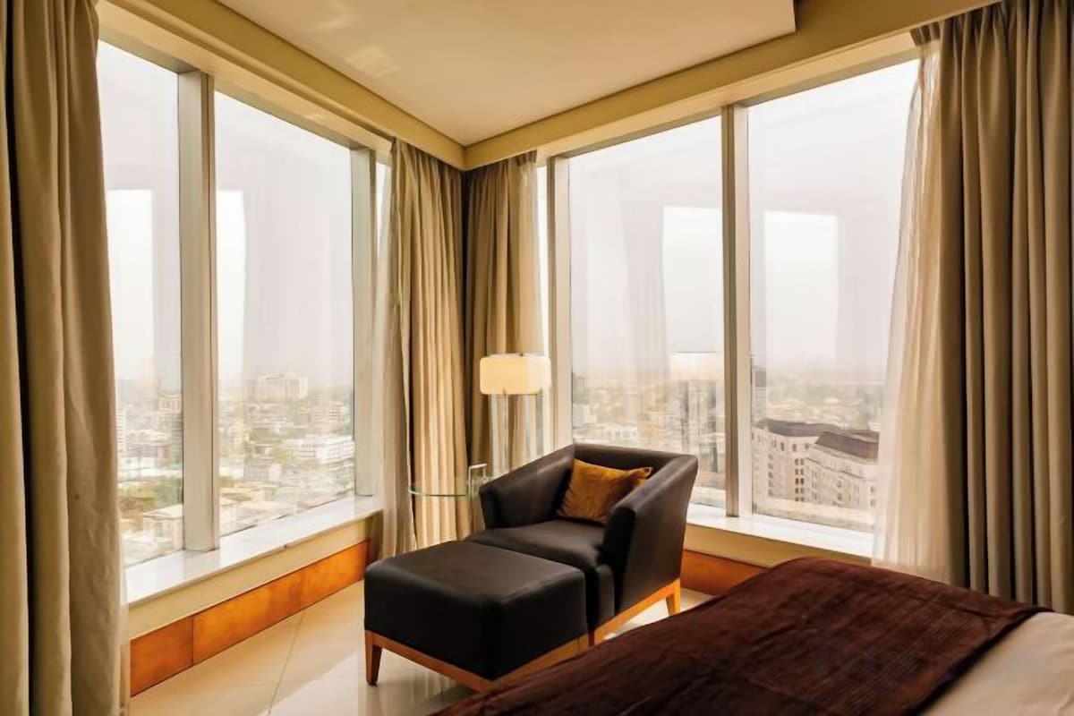 Una habitación de hotel con cama, una silla, una lámpara y ventanas grandes con cortinas.