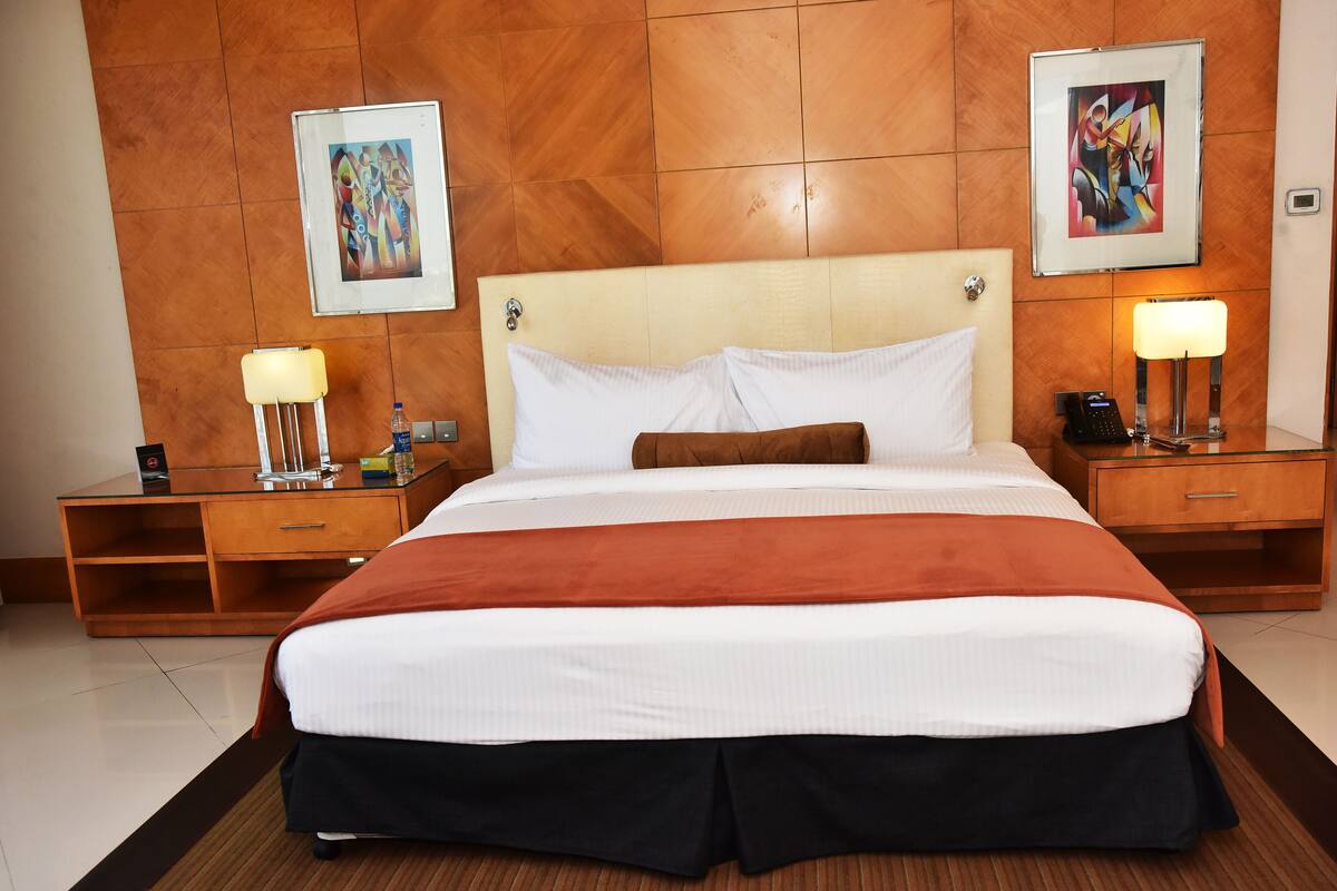 A hotel room with a large bed, two bedside tables, a nightstand, and two wall-mounted paintings.