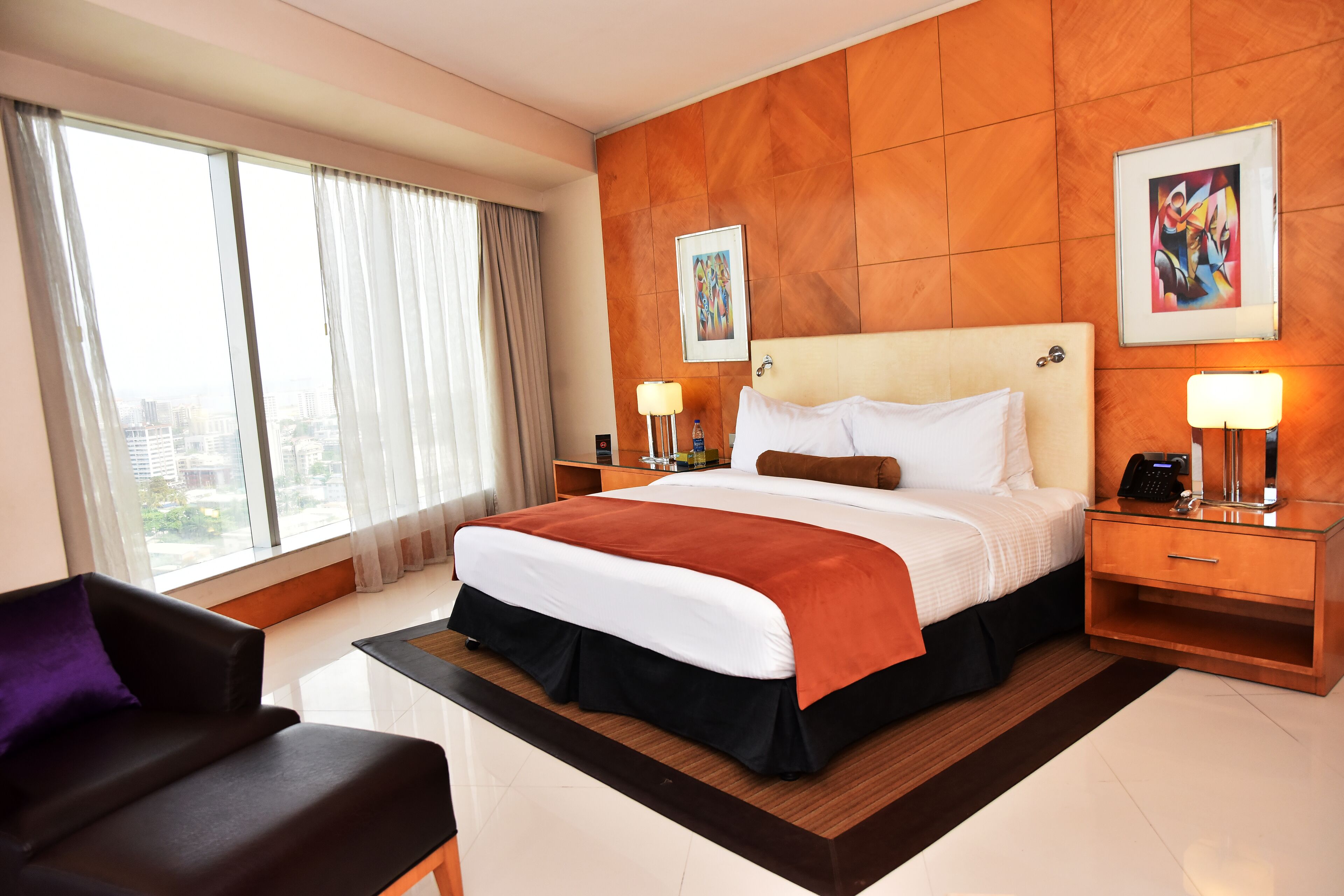 A modern hotel room with a large bed, a desk, a chair, and a view of the city through large windows.