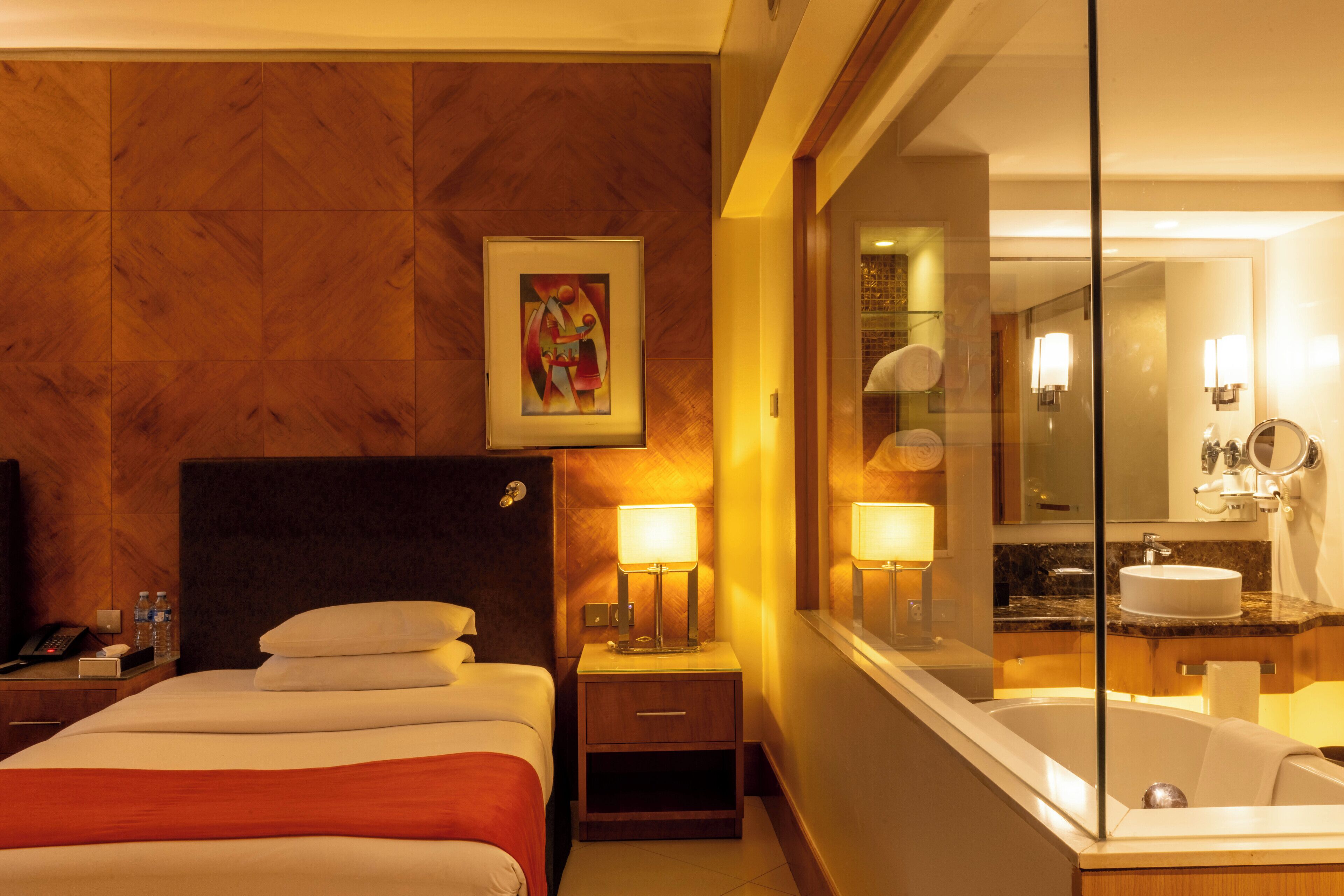 A hotel room with a bed, bedside table, lamp, and a large mirror reflecting the bathroom.