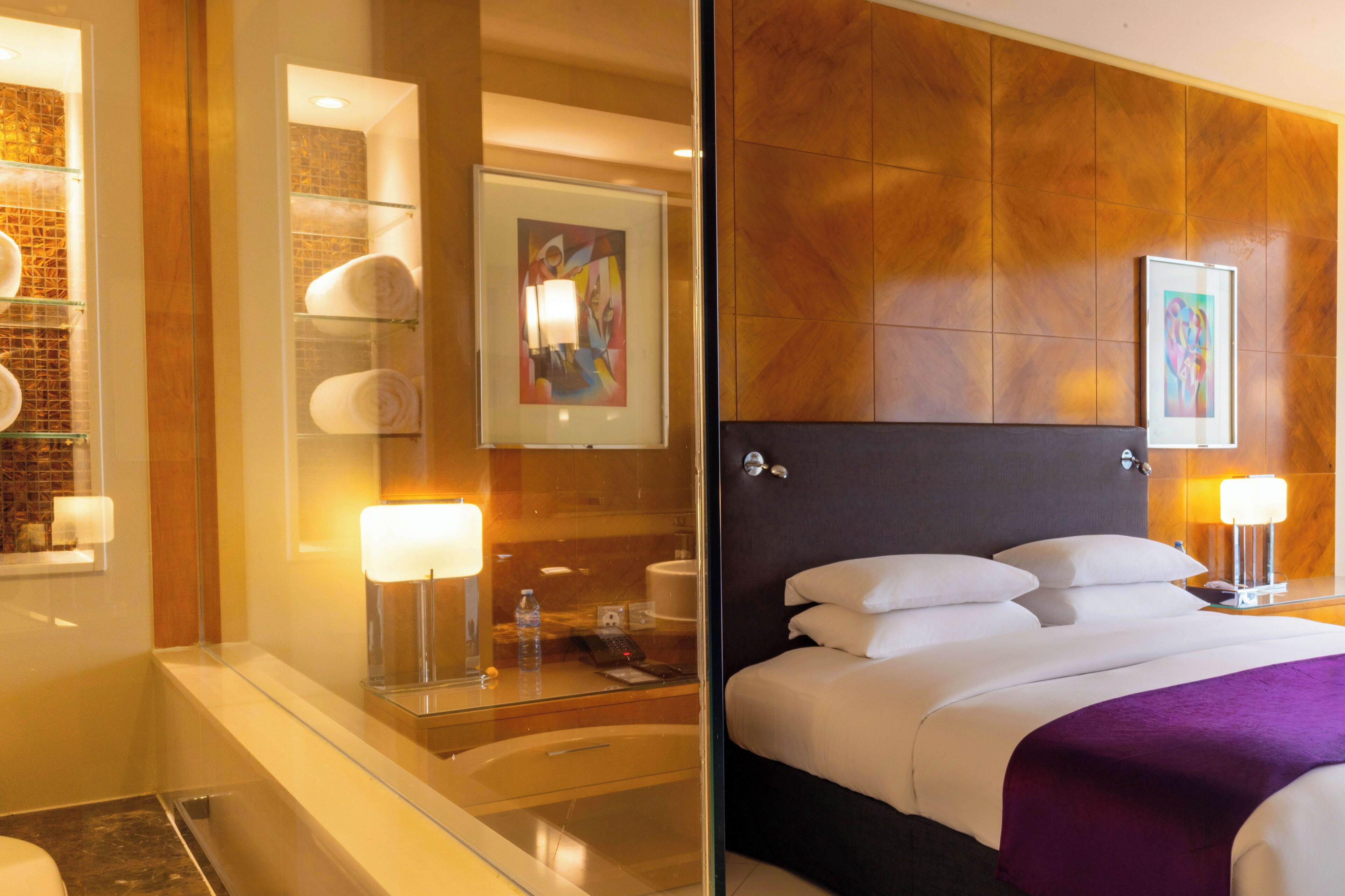 Habitación de hotel con cabina de ducha de vidrio, cama con ropa blanca y un plaid morado, y obras de arte en las paredes.