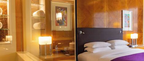 Habitación de hotel con cabina de ducha de vidrio, cama con ropa blanca y un plaid morado, y obras de arte en las paredes.