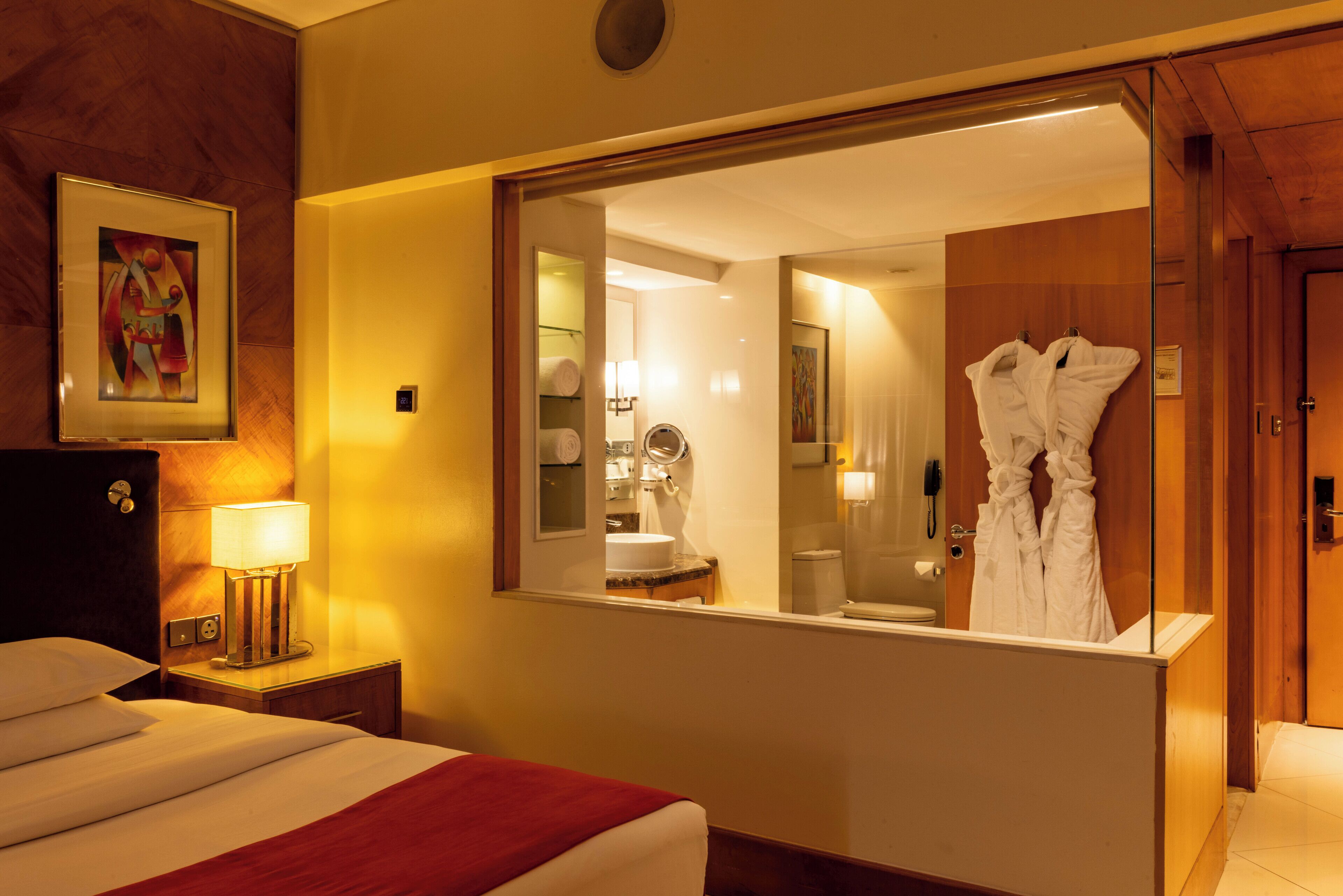 A hotel room with a bed, a bedside table with a lamp, a mirror reflecting the bathroom, and a robe hanging on the door.