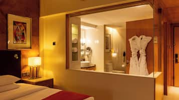 Habitación de hotel con cama, mesita de noche con lámpara, espejo que refleja el baño y bata colgada en la puerta.