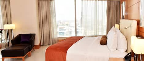A hotel room with a large bed, a sofa, a desk with a lamp, and a view of the city through large windows.