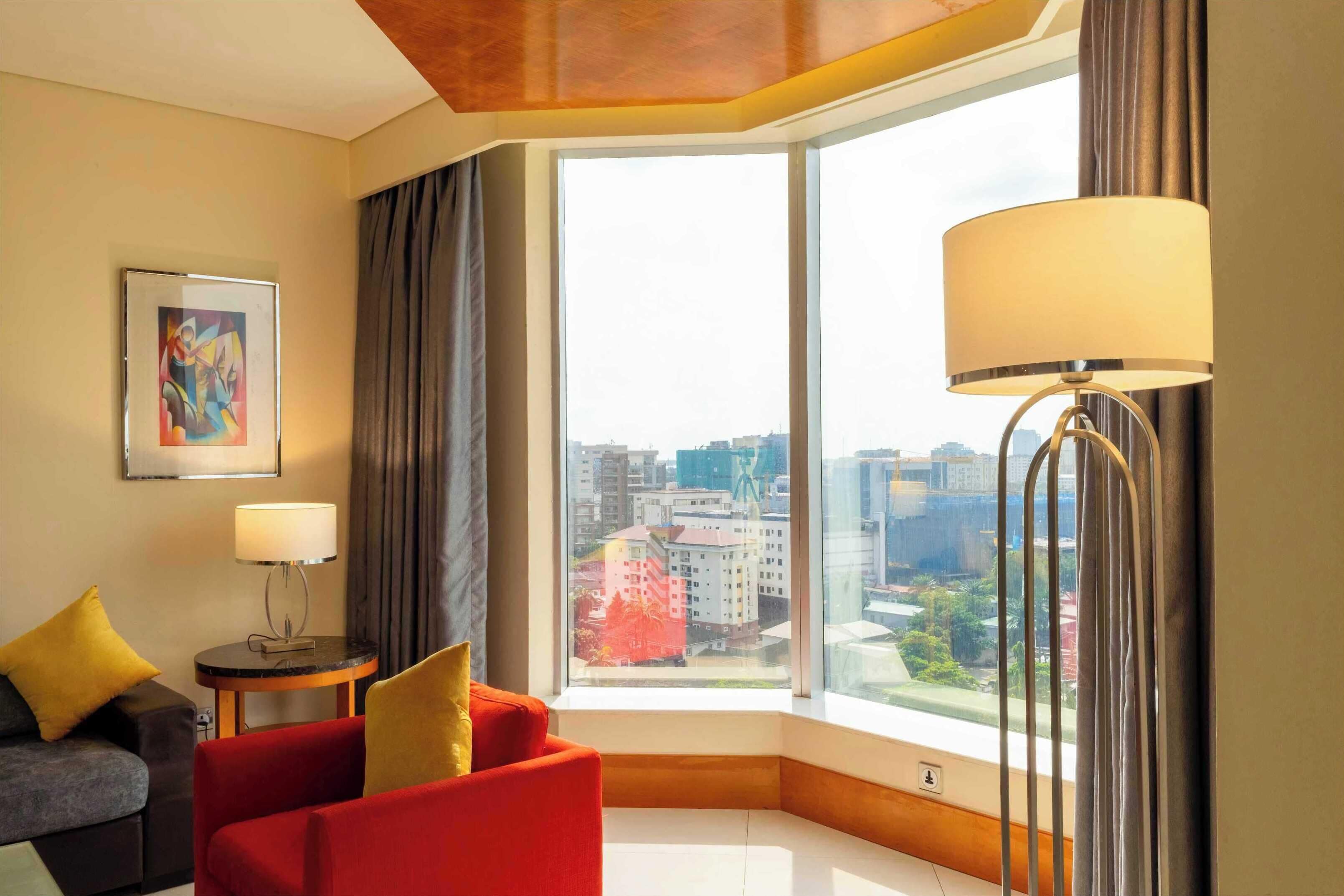 A hotel room with a large window offering a city view, a red sofa, a grey sofa, a small table with a lamp, and a painting on the wall.