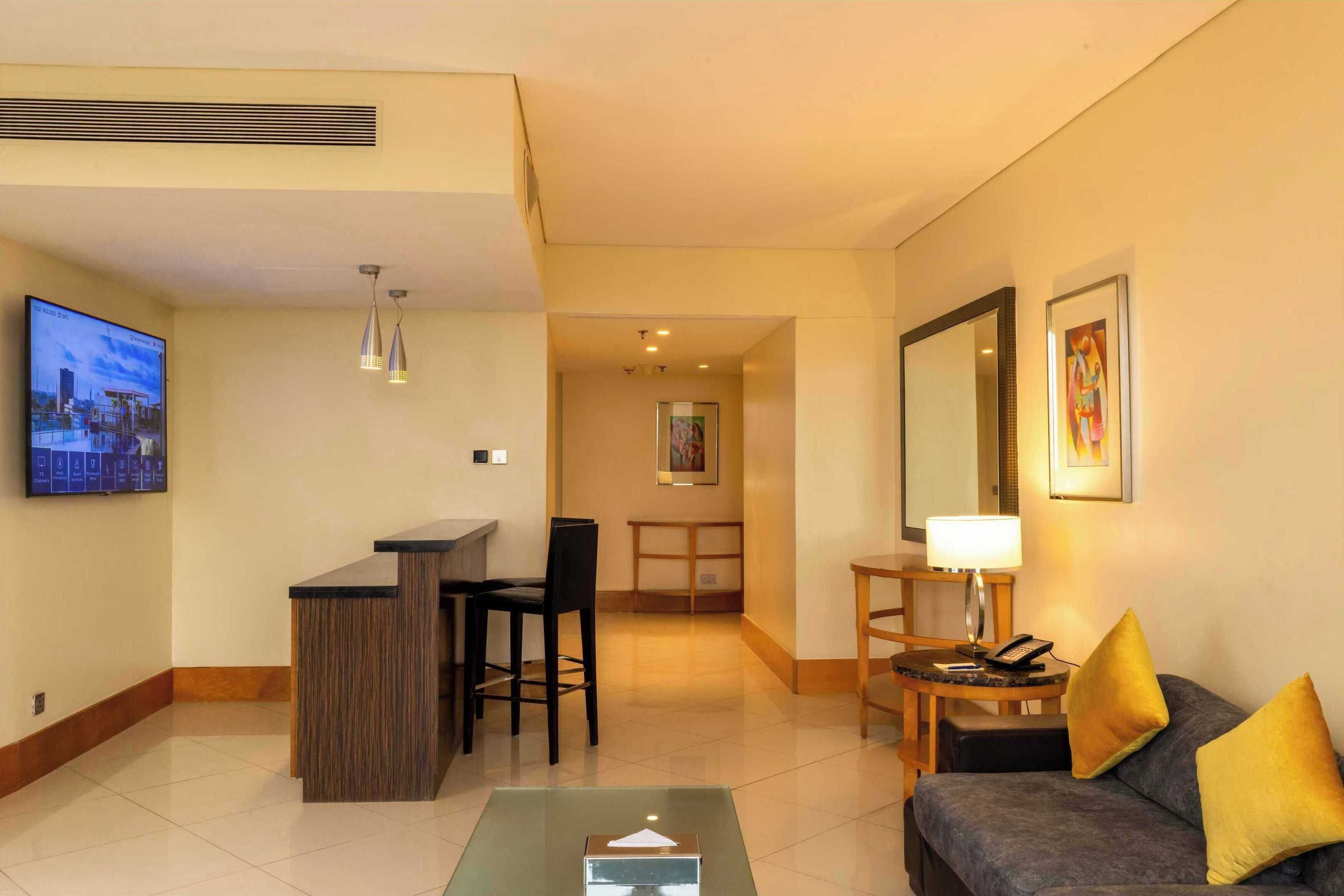 A hotel room with a reception desk, a seating area with a couch and chairs, a TV mounted on the wall, and a framed picture on the wall.