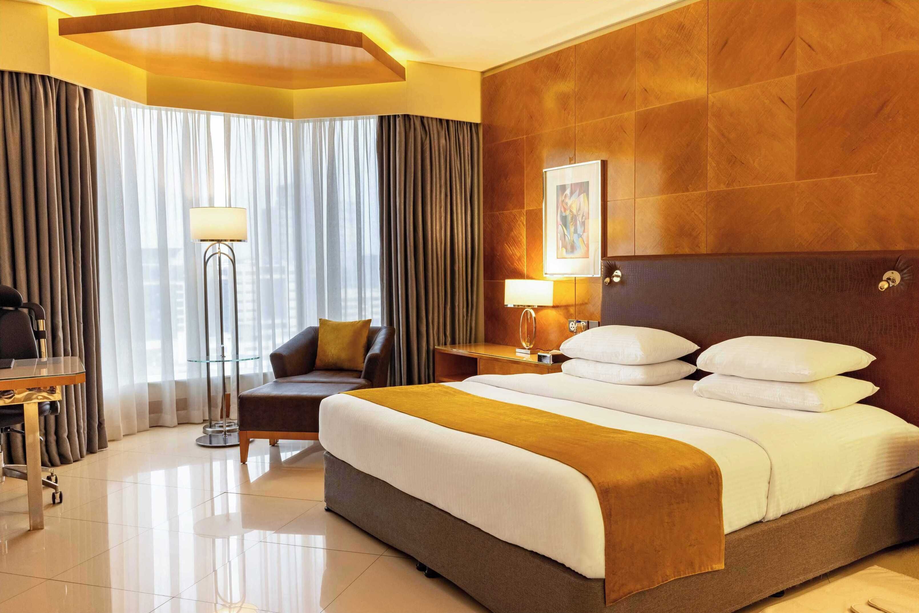 Une chambre d’hôtel avec un grand lit, un bureau, une chaise, une lampe sur pied et une fenêtre avec des rideaux.