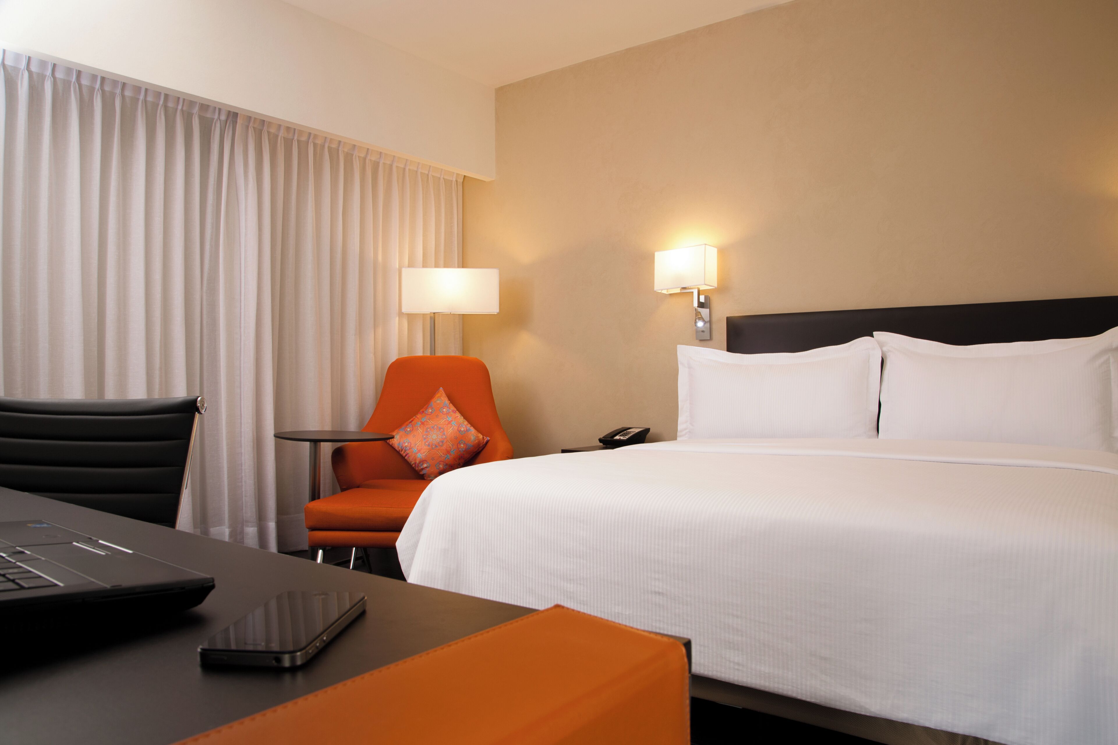 A hotel room with a bed, a desk with a laptop, a chair, and a nightstand with a lamp.