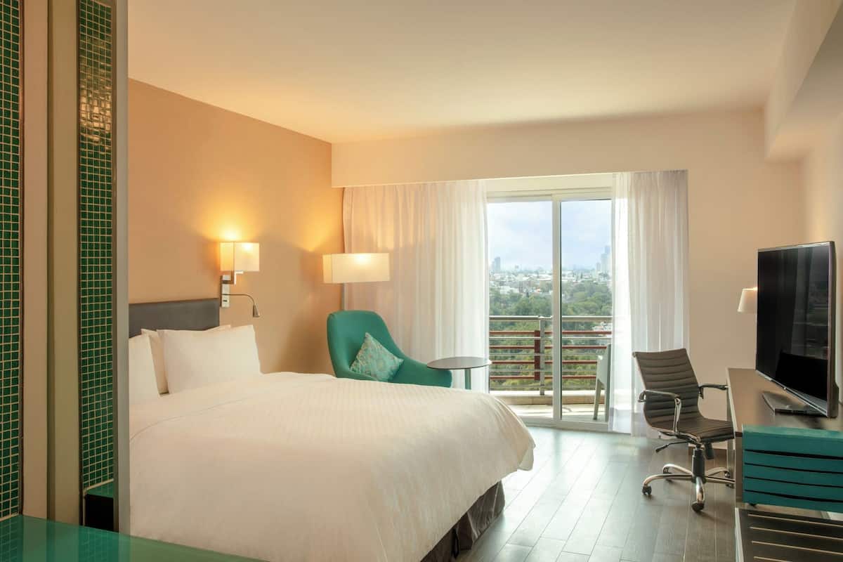 Hotelkamer met een bed, een bureau met een televisie, een stoel, een balkon met uitzicht en twee wandlampen.