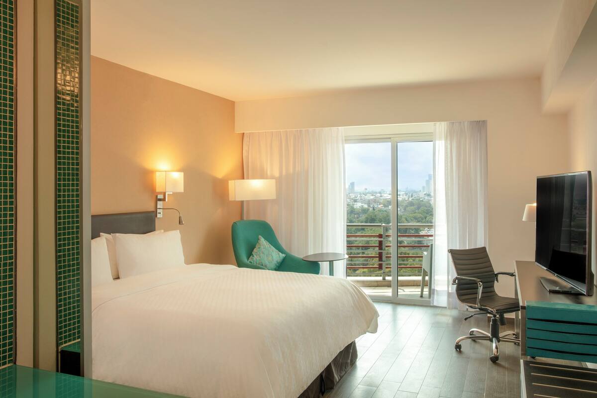 Een hotelkamer met een groot bed, twee stoelen, een bureau met een televisie en een balkon met uitzicht over de stad.