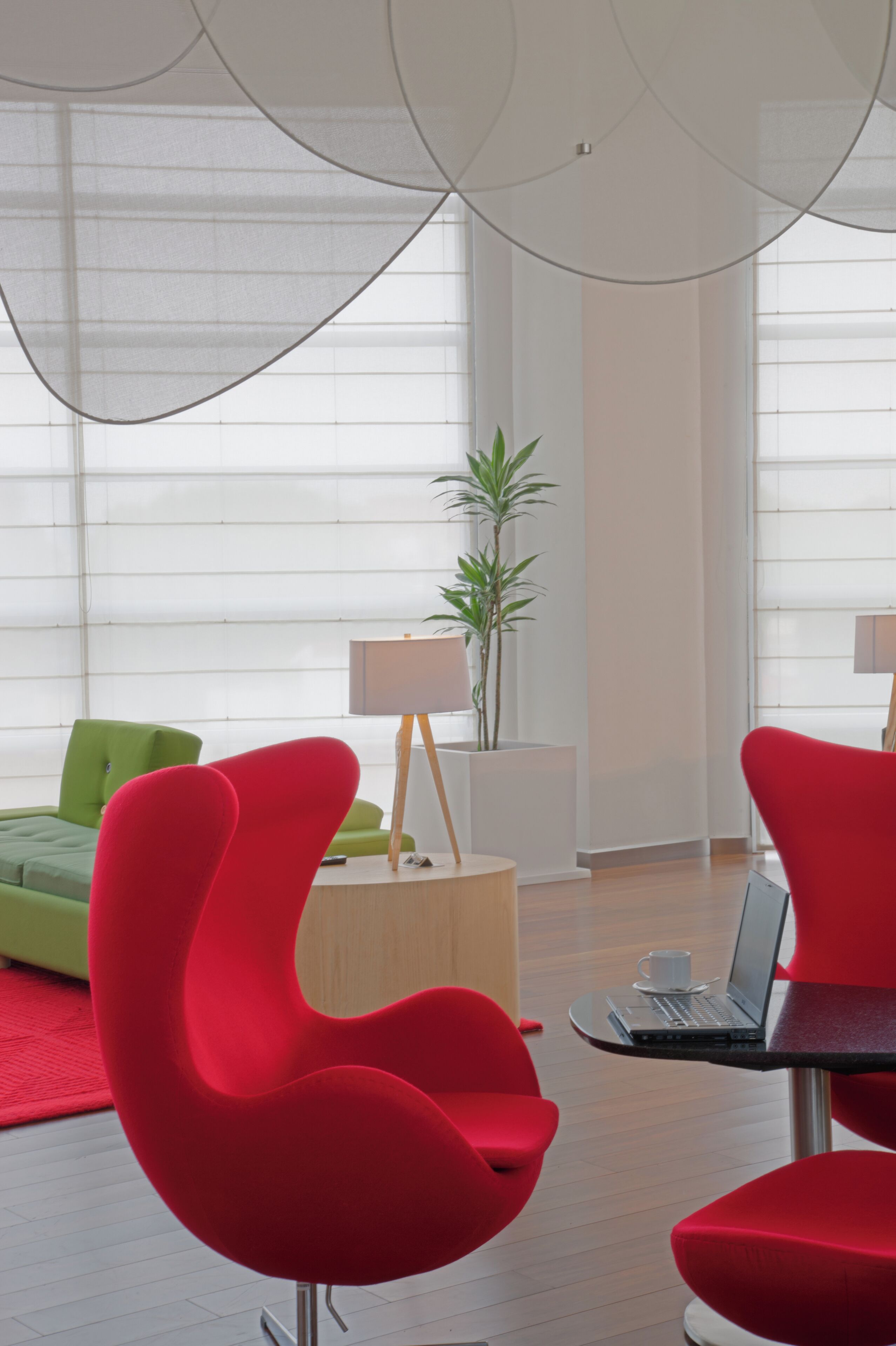 Una habitación moderna con sillas rojas, un sofá verde, una mesita de noche de madera con una lámpara, y un ordenador portátil sobre una pequeña mesa.