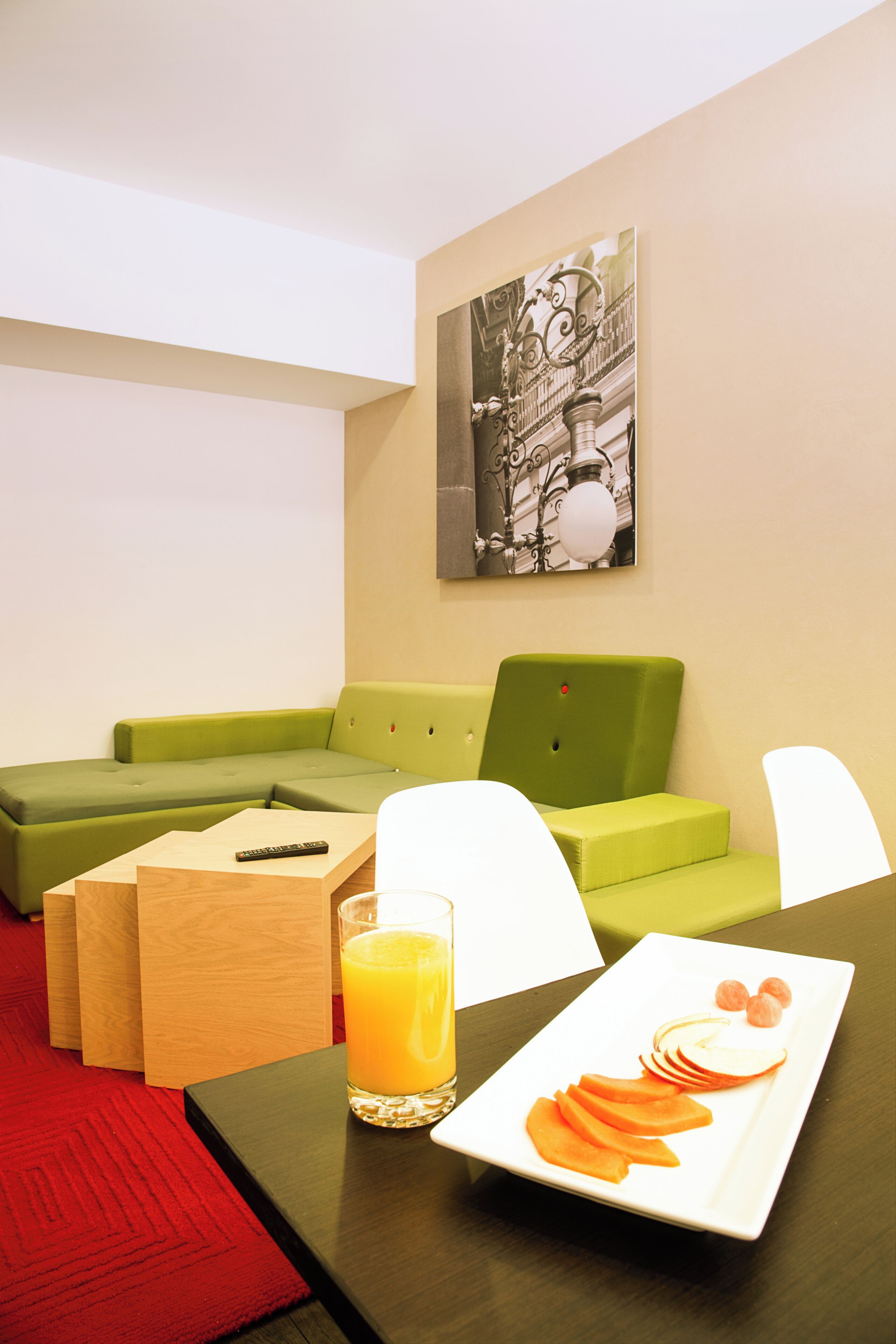 A modern hotel room with a green sofa, a small wooden table with a glass of orange juice, a white plate with fruit, and a black and white artwork on the wall.