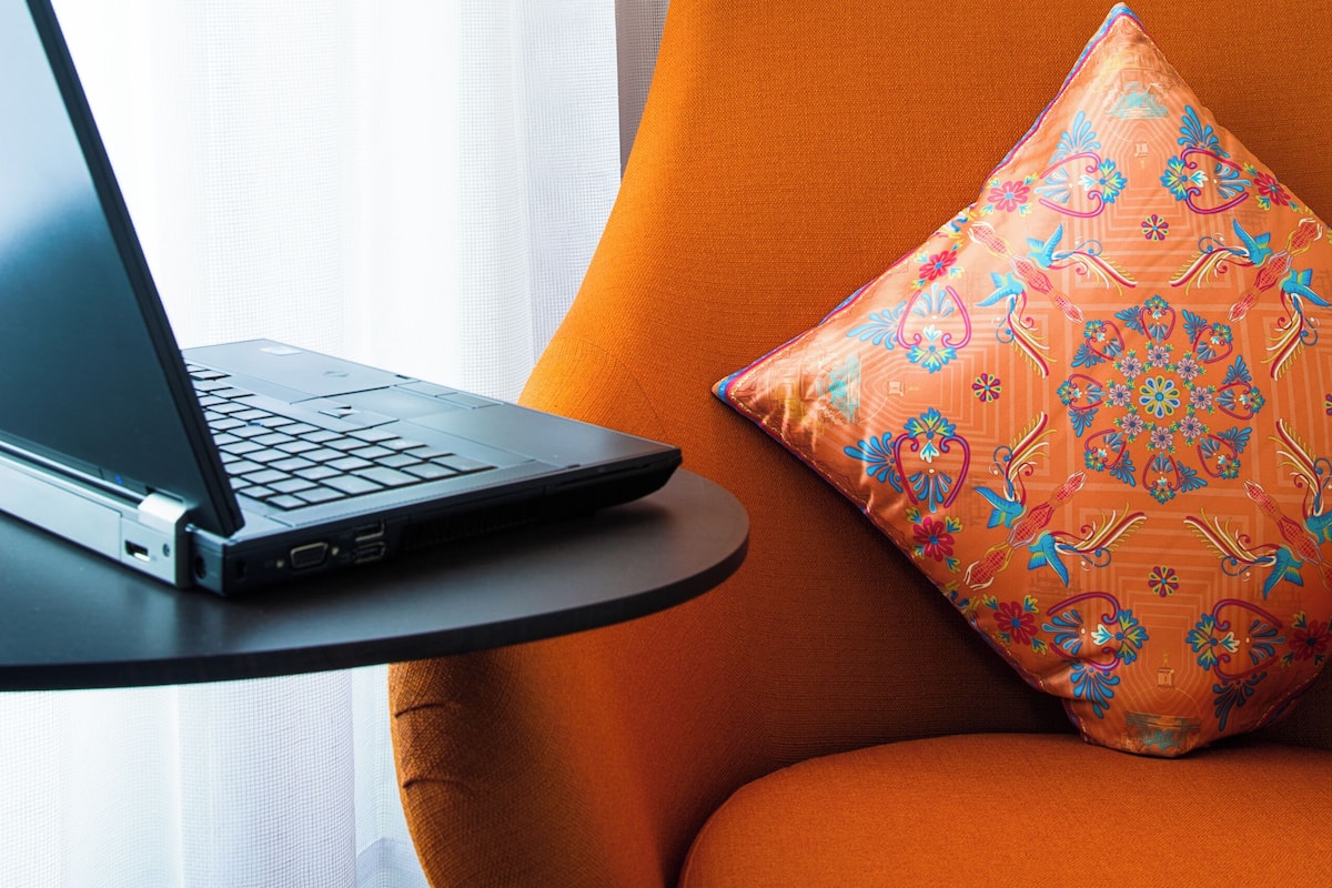 Een laptop op een klein tafeltje, naast een oranje stoel met een bloemenkussen.