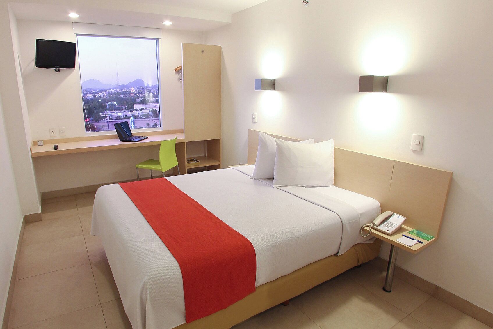 Una habitación de hotel con una cama, un escritorio con silla, un televisor y una ventana con vista a la ciudad.