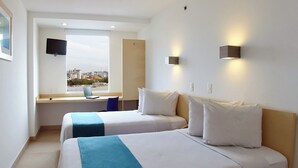 Habitación de hotel con dos camas, un escritorio con una computadora portátil, un televisor montado en la pared y una ventana con vistas a la ciudad.
