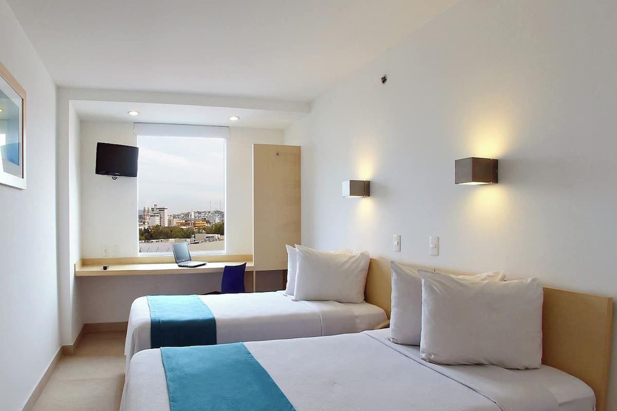 A hotel room with two beds, a desk with a laptop, a TV mounted on the wall, and a window with a city view.