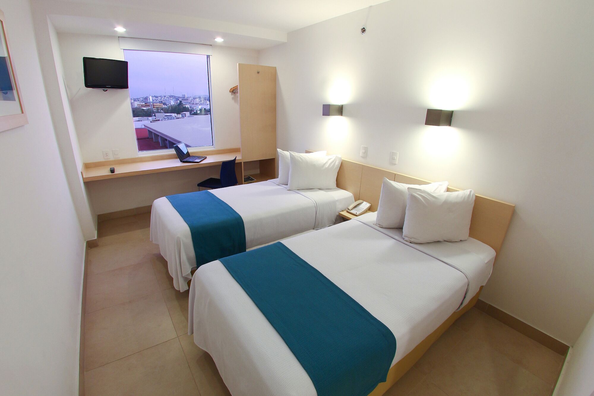 Habitación de hotel con dos camas, un escritorio con una computadora portátil, un teléfono y una ventana con vista a la ciudad.