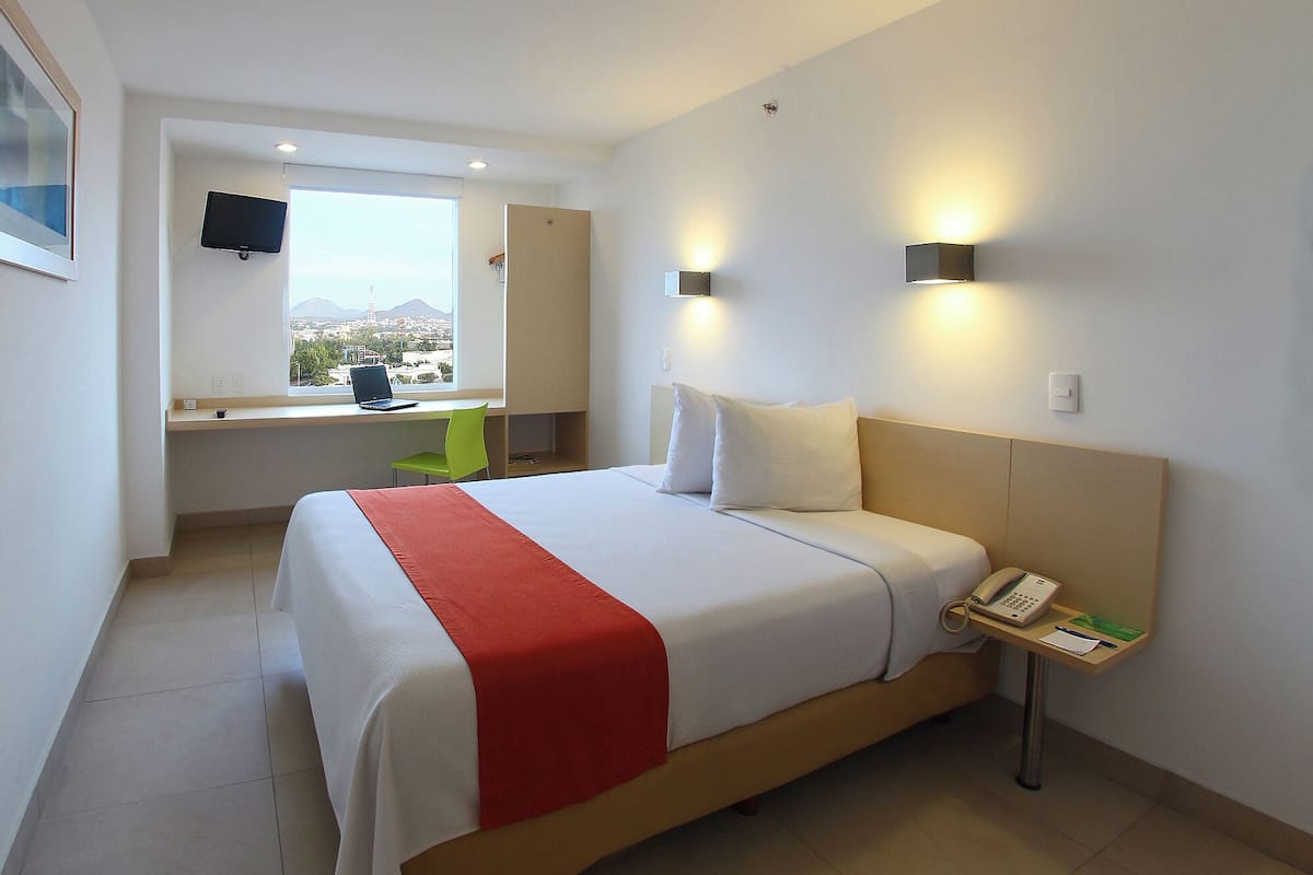 A hotel room with a bed, a desk with a laptop, a phone, and a view of a cityscape through a window.
