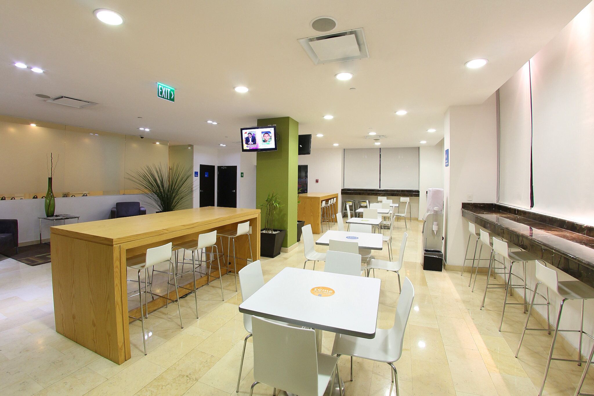 A modern indoor space with a reception desk, multiple tables and chairs, and a television screen.