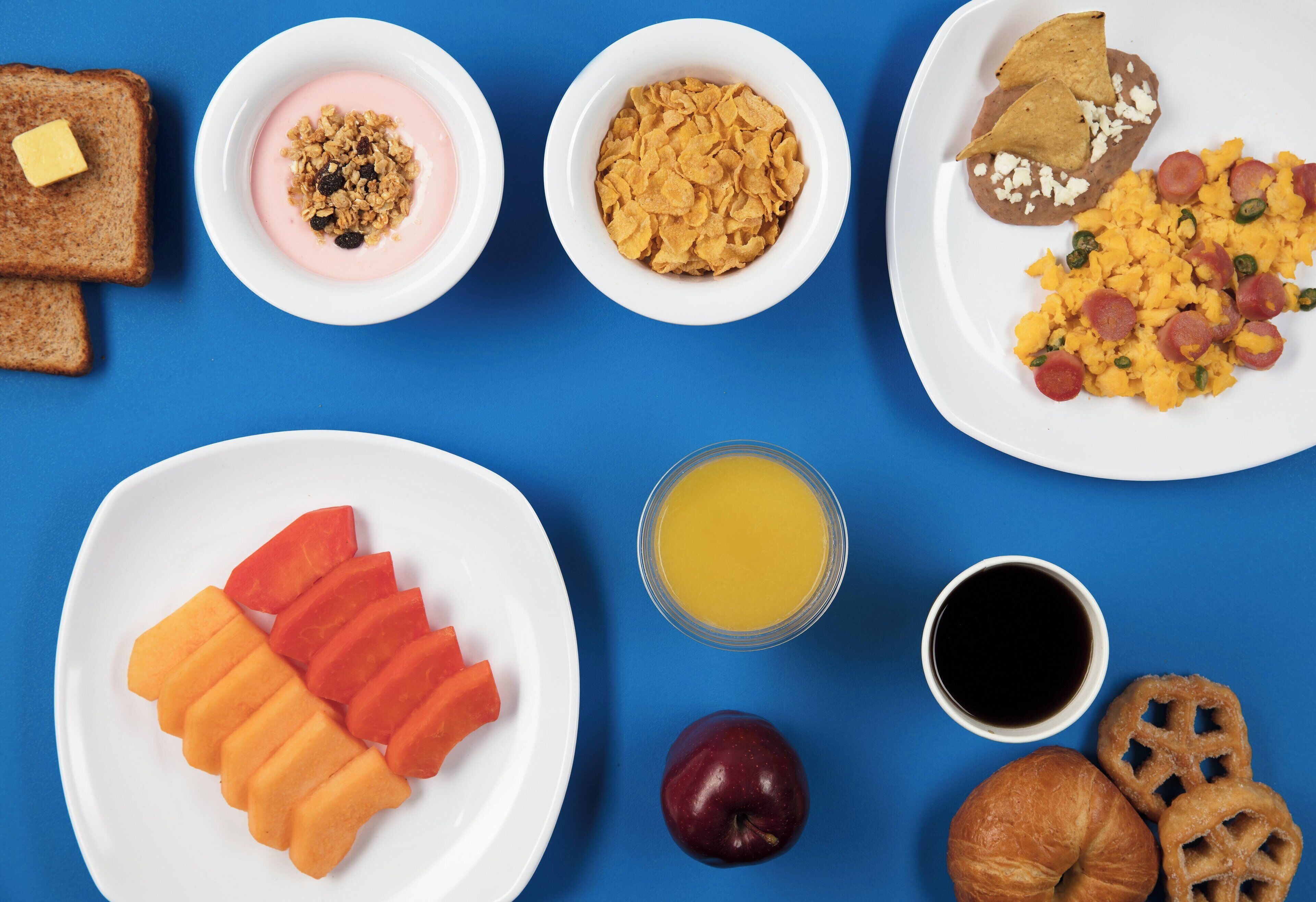 Un desayuno abundante con tostadas, yogur, cereales, frijoles, huevos revueltos, salchichas, jugo de naranja, café, rodajas de manzana y pasteles.