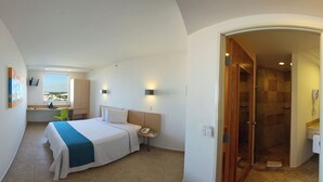 Une chambre d’hôtel comprenant un lit, une table de chevet et un bureau avec une chaise. La salle de bain est équipée d’une douche et d’un lavabo.