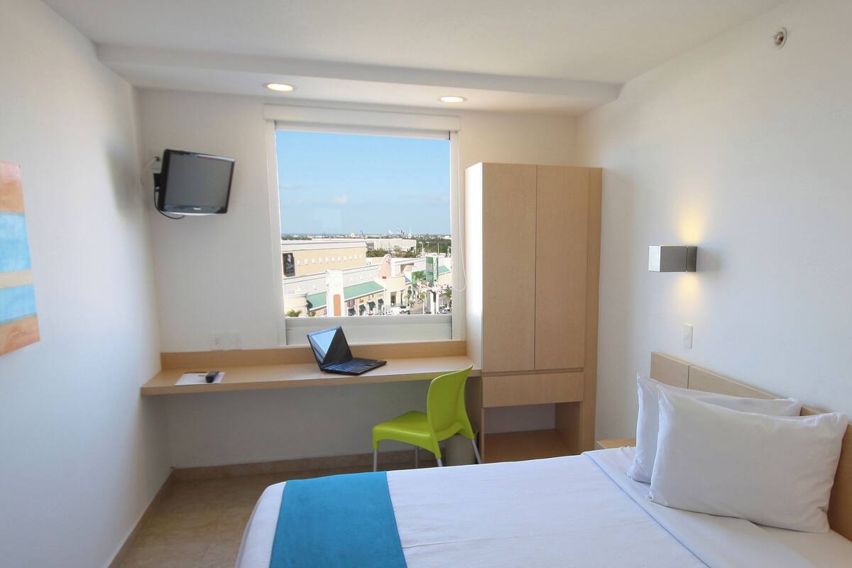 Una habitación de hotel con una cama, un escritorio con una computadora portátil, una silla verde y un televisor en la pared.