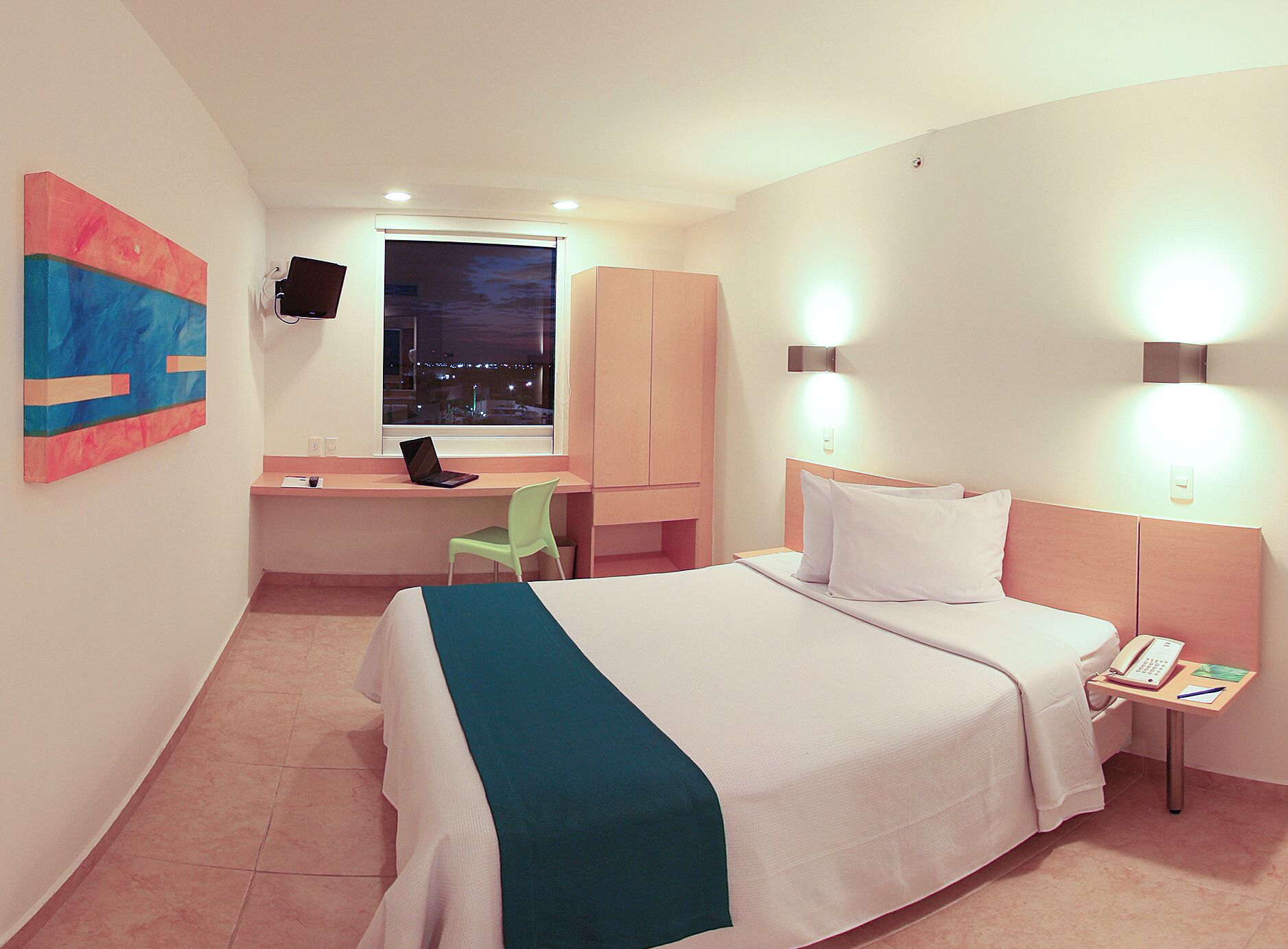 A hotel room with a bed, a desk with a laptop, a chair, a TV, and two wall-mounted lights.