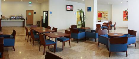 Un hall de hotel con varias zonas de estar, incluyendo mesas y sillas, una recepción y una pantalla de televisión.