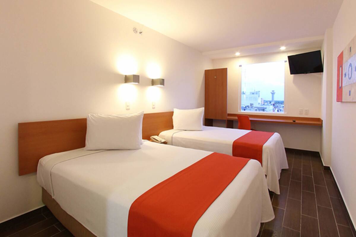 Habitación de hotel con dos camas, un televisor montado en la pared y una ventana con vista a la ciudad.