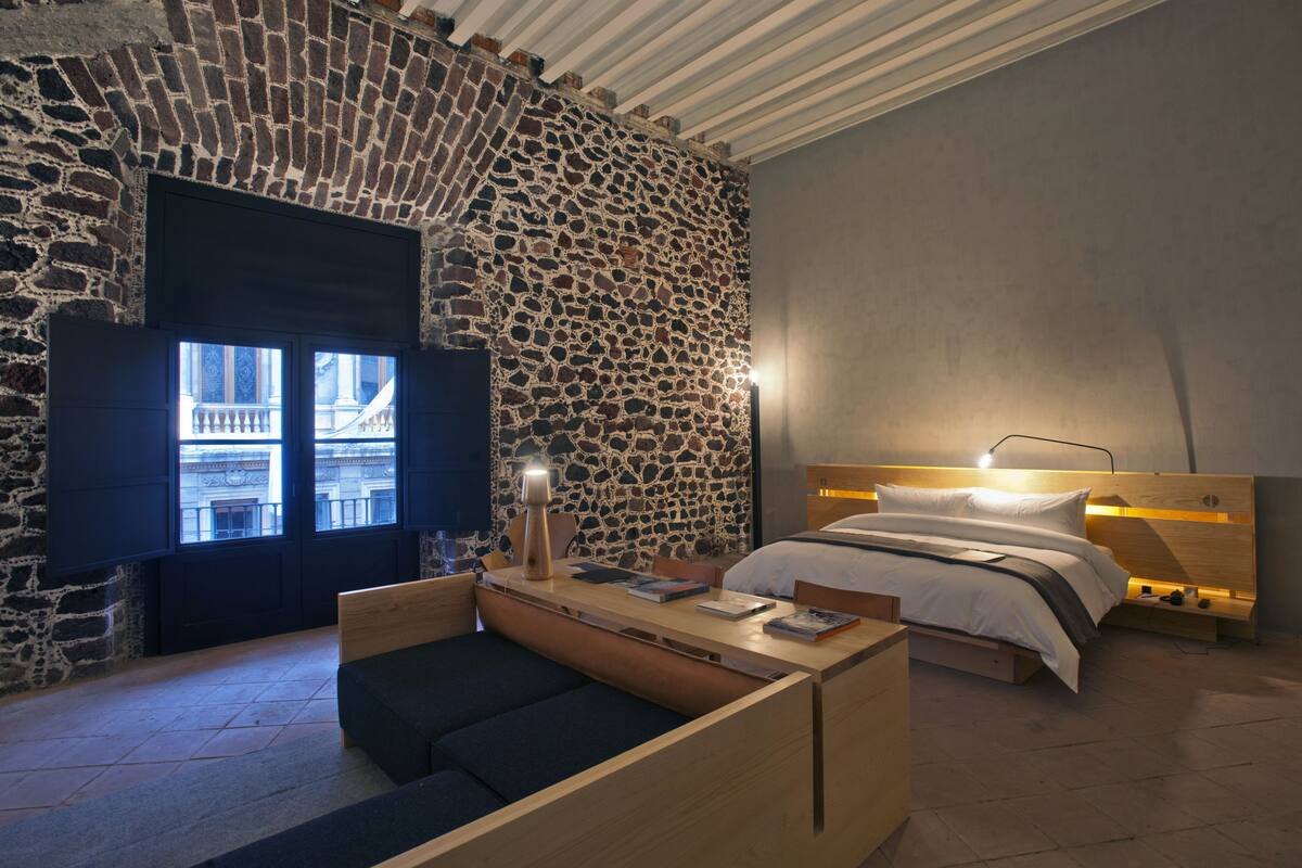 Une chambre avec un mur en pierre, un lit en bois, une table de chevet avec une lampe, et une vue sur un bâtiment par la fenêtre.