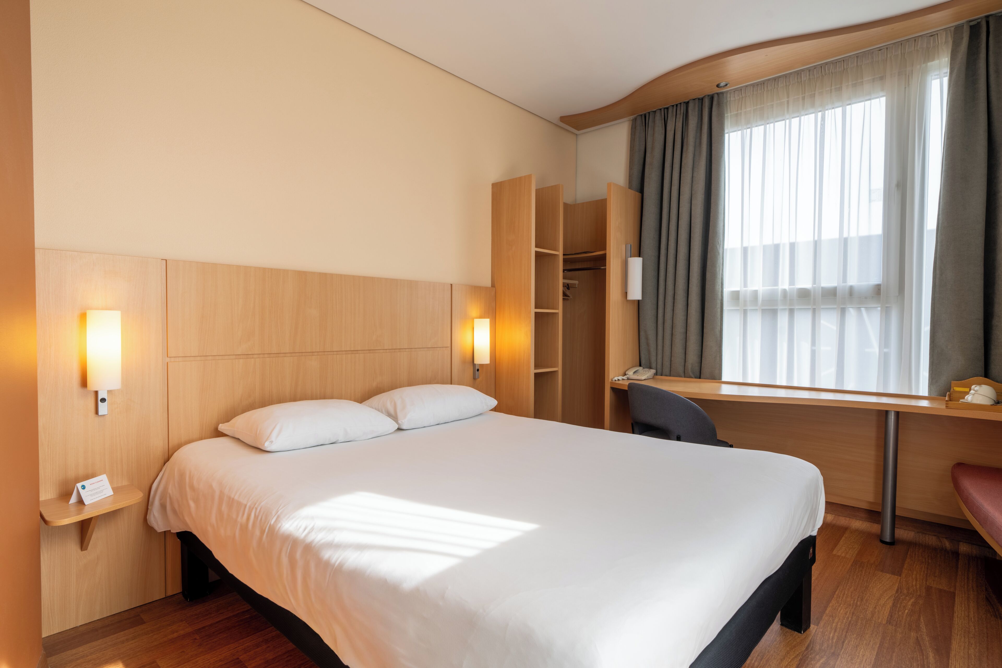 A hotel room with a large bed, a desk, a chair, a wardrobe, and a bedside table with a lamp.