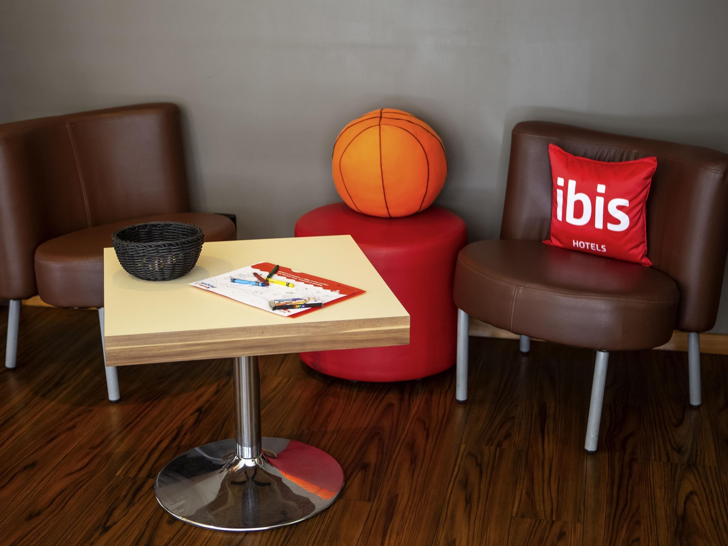 A hotel room with a wooden table, a red ottoman, two leather chairs, a basketball, a basket, and a magazine.