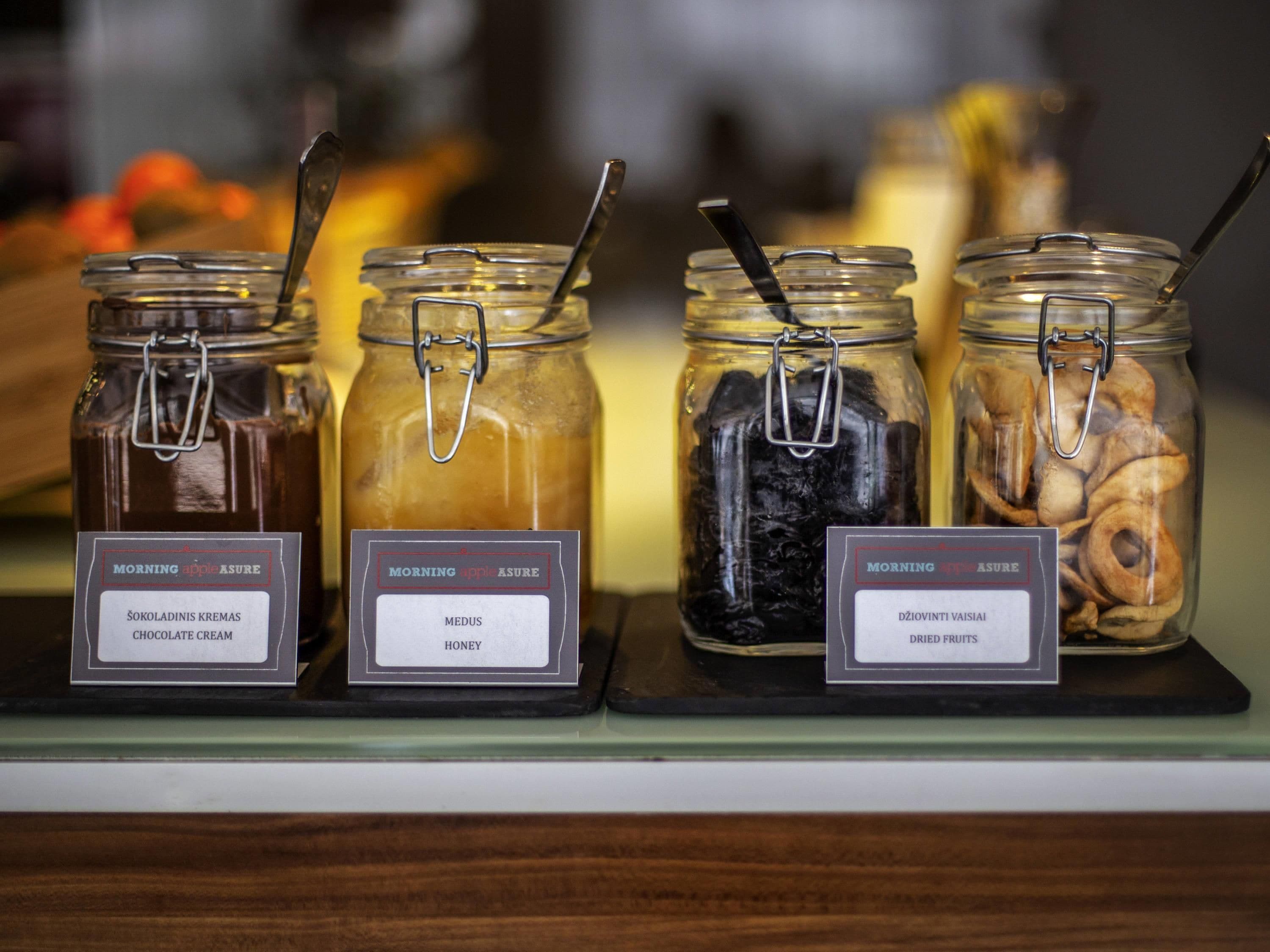 Cuatro frascos de vidrio con cierres metálicos, cada uno etiquetado con un elemento diferente: crema de chocolate, miel, frutas deshidratadas y un tercer artículo.